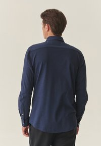 Jūras zilas garām piedurknēm plāksne, kas izgatavota no teksturētas auduma, ar norādītu apkakli un pogām aizdari. Gluda, pielāgota piegriezuma forma ar izliektiem malu galiem.