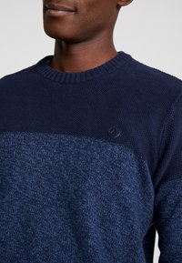 Navyblauer Strickpullover mit strukturiertem Oberteil und glattem Unterteil. Rundhalsausschnitt mit kleinem Markenlogo auf der Brust.