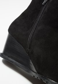 Bottine en suède noir avec un talon compensé, dotée d'une fermeture éclair latérale et d'une semelle en caoutchouc texturée. Le talon est angulaire et lisse.