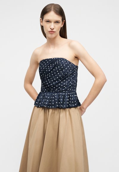 Γυναίκα με ίσια μαλλιά που φοράει μπλε σκούρο τοπ πουά χωρίς τιράντες με βολάν στο τελείωμα και μπεζ φούστα ψηλόμεση, με τα χέρια πίσω από την πλάτη.