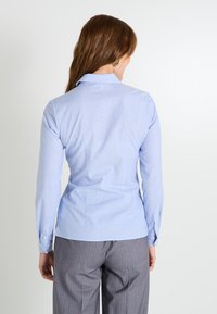 Camisa de botones de manga larga en azul claro con cuello puntiagudo, con un diseño ajustado y un sutil patrón de cuadros, combinada con pantalones de rayas grises.