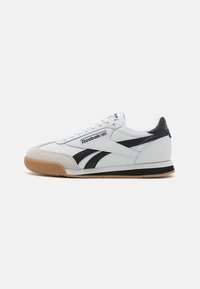 Sneakers en cuir blanc avec accents noirs, semelle en caoutchouc gomme, détails cousus et design rétro. Logo Reebok sur le côté.