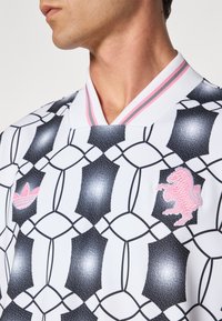 White shirt with black and pink geometric pattern, featuring a pink embroidered lion and a pink Adidas logo on the left chest.
