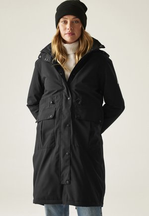 Kobieta ubrana w czarny, długi zimowy płaszcz z kapturem, biały golf, czarną czapkę i niebieskie jeansy, stojąca na jednolitym tle.