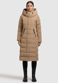 Dlouhý béžový péřový kabát s vysokým límcem, vodorovným prošíváním a zapínacími bočními kapsami, doplněný černými kozačkami a legínami.