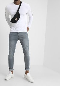 Camisa blanca de manga larga, jeans de mezclilla azul claro y zapatillas blancas. Un bolso cruzado negro descansa sobre el hombro con un parche de marca.
