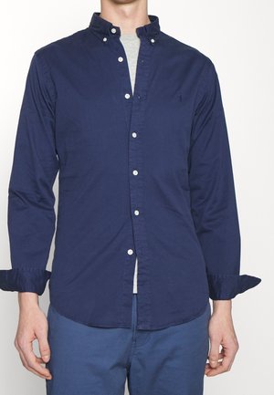 Man draagt een marineblauw knoopoverhemd met opgerolde mouwen over een lichtgrijs ondershirt en een blauwe broek tegen een effen achtergrond.