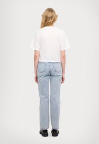 Kvinna sedd bakifrån i en vit kortärmad tröja, ljusblå rakpassform jeans och svarta platåskor mot en enhetlig bakgrund.