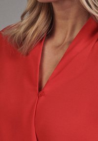 Blusa rossa con una consistenza liscia, caratterizzata da un colletto a V e un bottone al collo. Il tessuto sembra leggero e cade elegantemente.