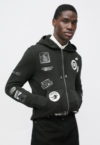 Μαύρο φούτερ με κουκούλα και φερμουάρ, με διάφορα μπαλώματα με λογότυπα. Το ύφασμα έχει λεία υφή, με ριμπ μανσέτες και τελείωμα.