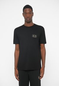 T-shirt de algodão preta com gola redonda e mangas curtas, com um pequeno logótipo dourado no lado esquerdo do peito. Textura suave.