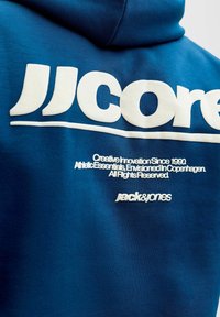 Blauwe hoodie met grote witte tekst "JJ core", kleinere witte tekst over creatieve innovatie en "jack & jones" branding eronder.