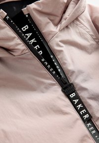 Rosa Kapuzenjacke mit glattem, leichtem Stoff und einem schwarzen Reißverschluss. Der Reißverschlussanhänger zeigt markanten weißen Text: "BAKER."