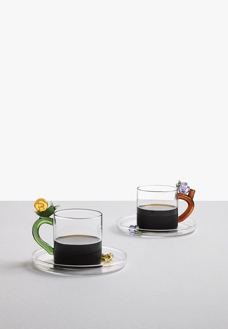 Dwie szklane filiżanki do kawy, jedna z zielonym uchwytem z żółtym kwiatem, druga z brązowym uchwytem i fioletowym kwiatem. Przezroczyste spodeczki.