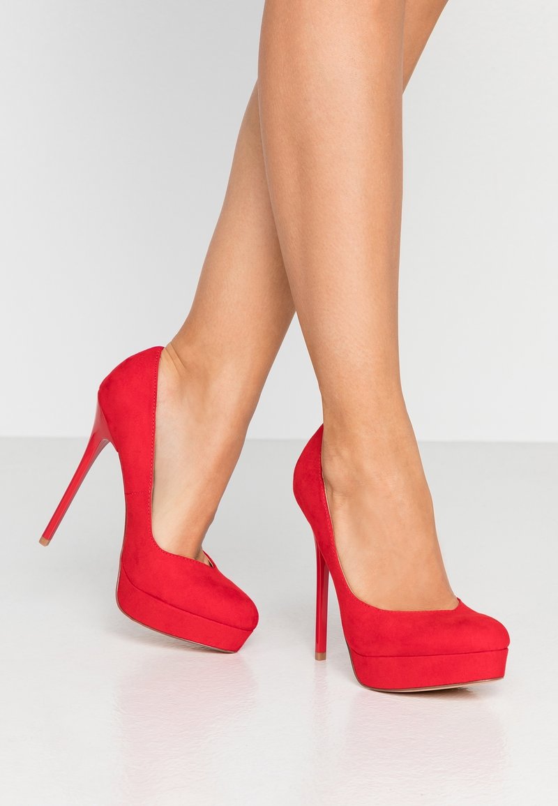 Escarpins en daim rouge à talons hauts avec un design à plateforme, présentant une silhouette élégante, des côtés ouverts et des talons aiguilles rouges brillants.