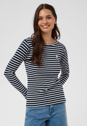Femme aux longs cheveux bruns souriante, portant un tee-shirt à manches longues rayé bleu marine et blanc avec un petit détail en forme de cœur et un jean bleu sur fond uni.