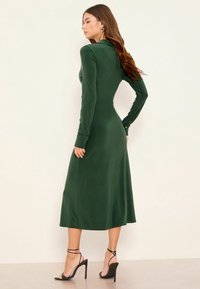 Rochie verde închis cu mâneci lungi, corset strâns pe corp și fustă fluidă. Prezintă nasturi pe mâneci și un material neted, elastic.