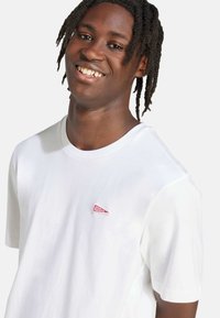 Biely bavlnený tričko s okrúhlym výstrihom a krátkymi rukávmi, s červenou nášivkou loga na hrudi. Hladká textúra a voľný strih.