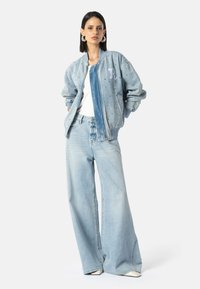 Žena stojící s rukama v kapsách, oblečená v světle modré džínové bomber bundě se cvočky, bílém topu, širokých světle modrých džínách a bílých botách.