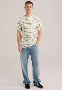 Tricou cu mânecă scurtă, cu un fundal alb și dungi neregulate verzi și bej; asortat cu blugi albaștri deschiși și adidași negri.