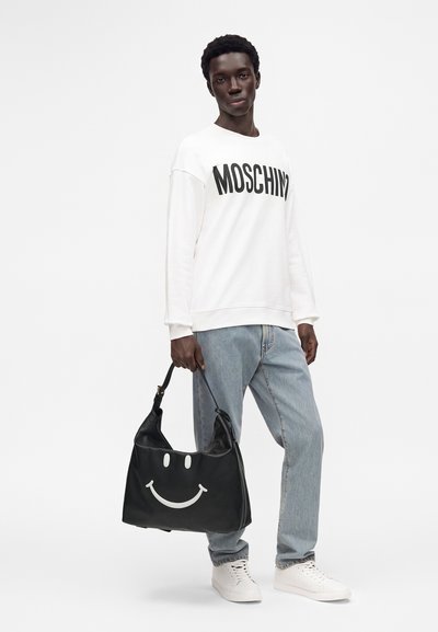 Homme portant un sweat-shirt blanc Moschino, un jean bleu clair, des baskets blanches, tenant un sac à main noir avec un grand visage souriant blanc.