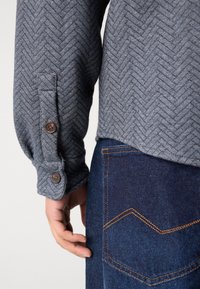 Gray herringbone-patterned shirt with brown buttons at the cuff, paired with dark denim featuring a contrasting orange stitch pocket detail.