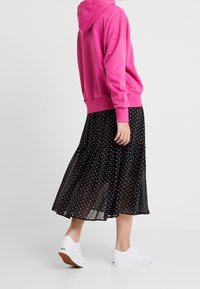 Rosa hoodies, svart kjol med färgglatt hjärtmotiv och vita sneakers. Kjolen är lätt och något genomskinlig; hoodien har en känguruficka.