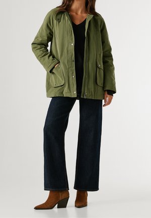 Vrouw met een olijfgroene jas, zwart topje, donkere wijde jeans en bruine enkellaarzen met hak, staand tegen een effen achtergrond.