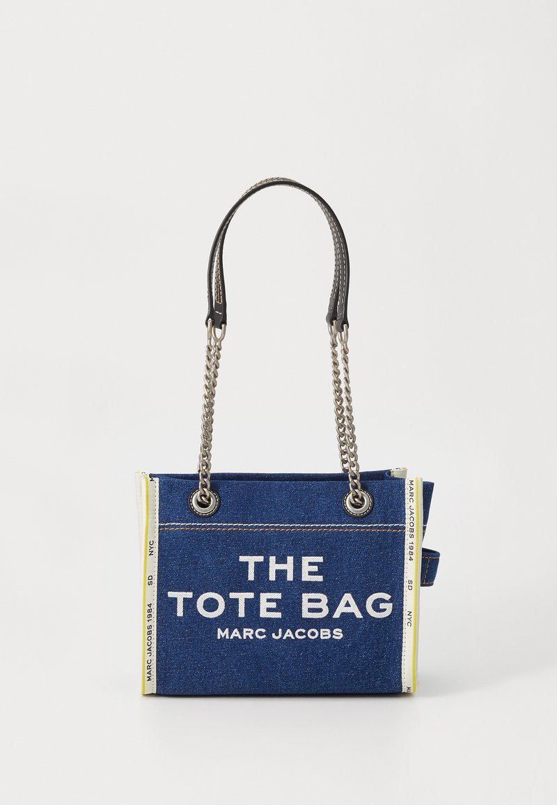 Saco tote de denim azul com alças de corrente prateada, acabamento branco nas laterais com texto e "THE TOTE BAG MARC JACOBS" impresso na frente.