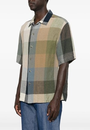 Uomo che indossa una camicia a maniche corte con grandi blocchi di colore smorzato in beige, verde, blu e marrone, abbinata a pantaloni blu.