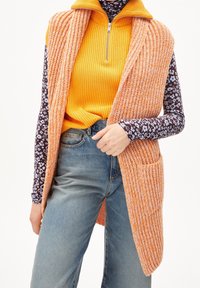 Pletená oranžová vesta s otevřeným předním dílem a dvěma kapsami, přesažená přes žebrovaný žlutý top a tmavě květinovou košili s dlouhým rukávem, v kombinaci s modrými džíny.