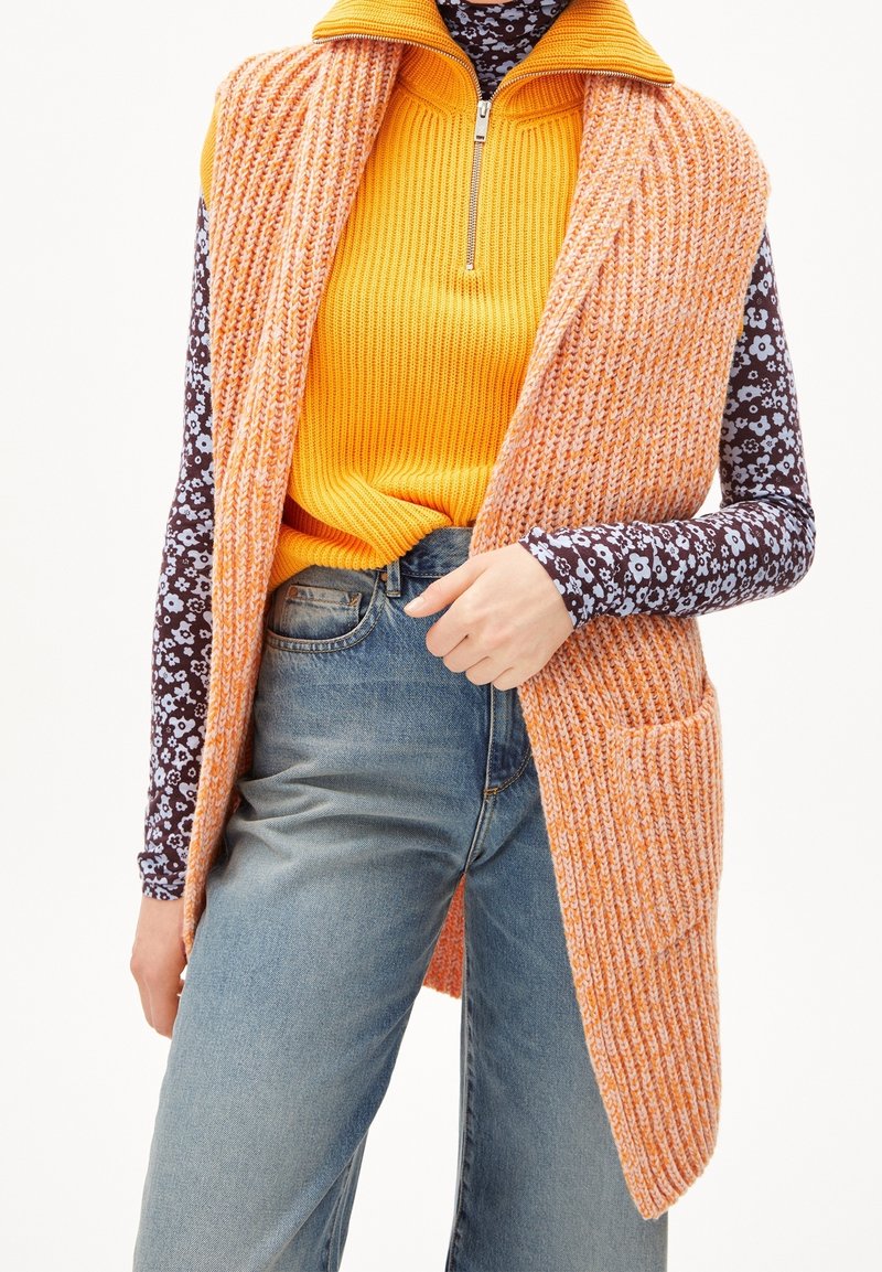 Pletená oranžová vesta s otevřeným předním dílem a dvěma kapsami, přesažená přes žebrovaný žlutý top a tmavě květinovou košili s dlouhým rukávem, v kombinaci s modrými džíny.