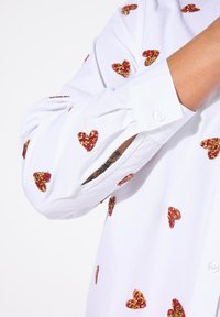 Camicia bianca con motivi a cuore in glicine e oro glitterati. Presenta un polsino con bottoni e una vestibilità rilassata. Il tessuto è liscio con una leggera lucentezza.