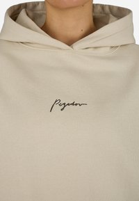 Μπεζ φούτερ από μαλακό ύφασμα με μπροστινή τσέπη καγκουρό. Διαθέτει κουκούλα με κορδόνι και μαύρο κεντημένο λογότυπο "Pegador" στο στήθος.