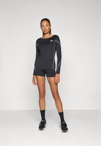 Svart atletisk långärmad tröja med grå sömdetaljer, ihopparad med svarta shorts och strumpor. Modellen står i svarta sneakers.