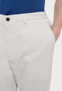 Pantaloni grigi chiari con una texture liscia, dotati di passanti per cintura, chiusura con bottone frontale e tasche laterali. Materiale in cotone.