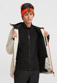 Jeune personne portant un bonnet orange avec des lunettes de protection, tenant un manteau d'hiver beige ouvert pour révéler un polaire noir zippé en dessous.