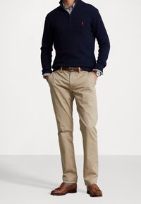 Marineblauer Pullover mit Rundhalsausschnitt und halbem Reißverschluss, beige Chinos, braune Leder-Loafers und Armbanduhr. Sichtbare gestreifte Hemdmanschetten. Einfaches, klassisches Design.