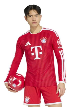 Junger Mann trägt rotes FC Bayern München Fußballtrikot und Shorts, hält einen passenden roten Fußball in der linken Hand und steht vor weißem Hintergrund.