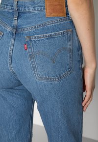 Blue denim jeans with a high waist, stitched back pockets, tan leather patch, and a red label near the right pocket. Distressed texture visible.