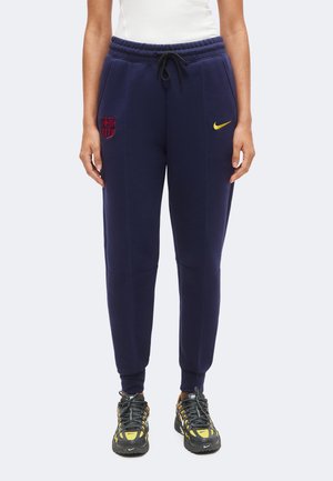 Persona que lleva pantalones deportivos azul marino con los logotipos del FC Barcelona y de Nike en amarillo, combinados con zapatillas Nike negras y amarillas.