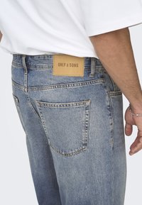Jean en denim bleu clair avec une coupe droite, doté de deux poches arrière et d'une étiquette en cuir marron avec l'inscription "ONLY & SONS" cousue sur la ceinture.