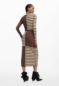 Gestreiftes Langarmkleid aus braunem, cremefarbenem und schwarzem Rippstoff. Hat eine figurbetonte Silhouette und einen asymmetrischen Saum; kombiniert mit schwarzen Stiefeletten mit Absatz.