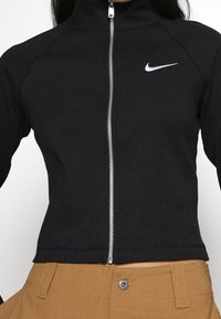 Black ribbed zip-up jacket with high collar and silver zipper. Features a white Nike logo on the chest. Tight fit and long sleeves.