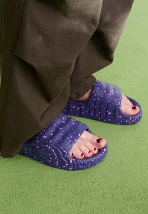 Pieds avec du vernis à ongles rouge foncé portant des sandales violettes avec des taches roses sur une surface verte, partiellement couverts par un pantalon marron.