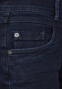 Primer plano de unos jeans de mezclilla azul oscuro que muestran el bolsillo delantero con el bolsillo para monedas y los remaches de metal con el grabado de la marca.