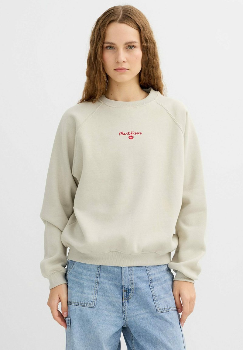 Sudadera beige clara con cuello redondo y mangas raglán. Bordado rojo "Plantlover" en el pecho. Tejido suave y texturizado.