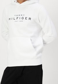 Biała bluza z kapturem wykonana z miękkiego materiału, z kieszenią kangurką i kapturkiem ze sznurkiem. Na przodzie znajduje się napis "TOMMY HILFIGER EST. 1985" w granatowym kolorze.