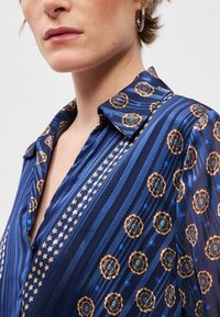 Mėlynas raštuotas marškinis su gėlėmis ir žvaigždžių motyvais, pagamintas iš lygaus audinio, su apykakle ir mygtukų užsegimu, pusiau permatomas.