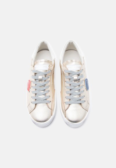 Αθλητικά παπούτσια από δερμάτινο μεταλλικό χρυσό με γκρι κορδόνια· διαθέτουν κόκκινες και μπλε λεπτομέρειες στην πλευρά· στρογγυλεμένη μύτη και λευκή καουτσούκ σόλα.